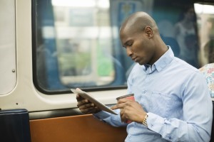 African man commuting on subway while working with digital tablet. He has a coffee in his hand.